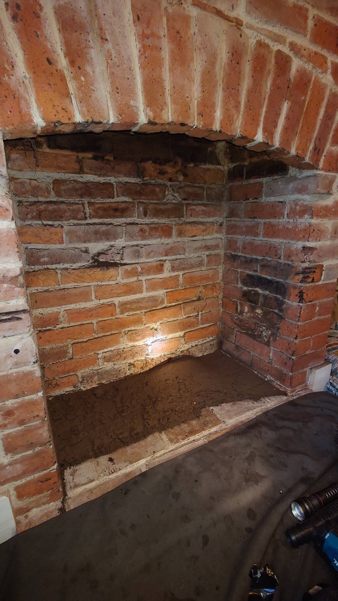 Inside a brick inglenook fireplace lit by a work lamp, with soot on the hearth
